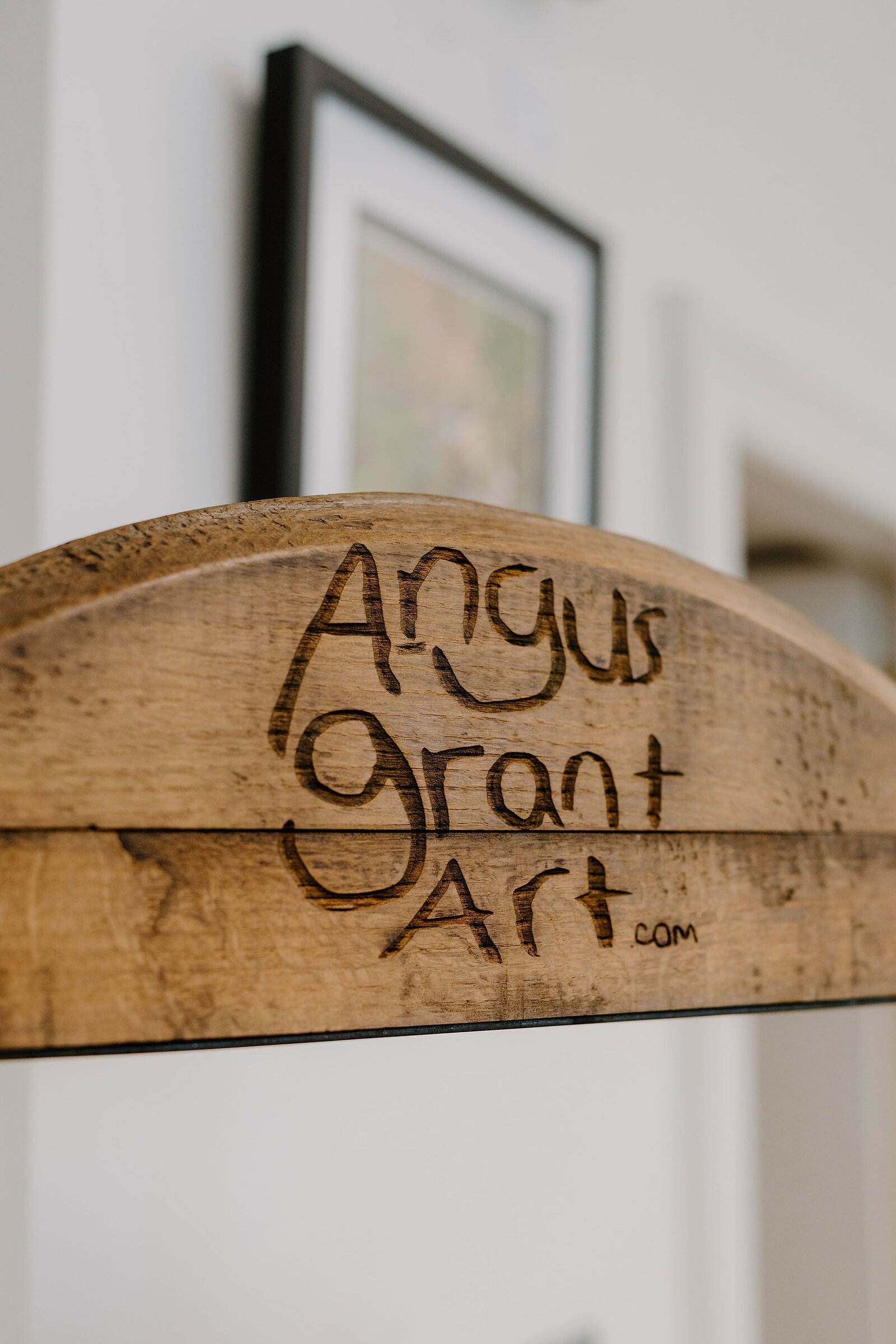 A wood-burned sign reads Angus Grant Art dot com, a framed picture blurred behind it.