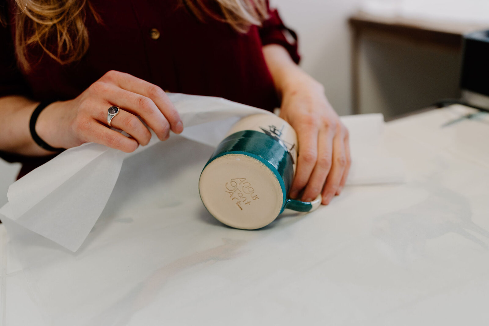 A woman's hands wrap a glazed mug in white tissue paper on a counter, rings visible on her fingers.
