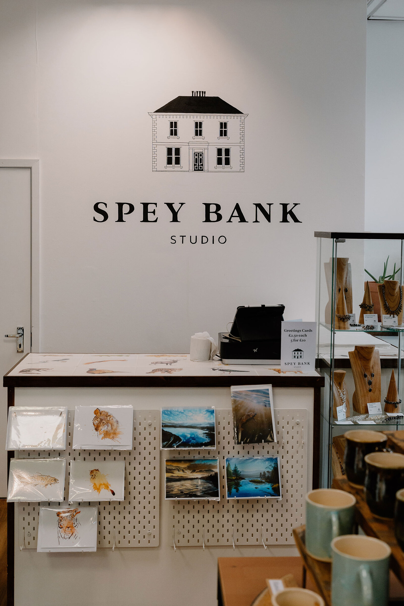 A shop display for Spey Bank Studio holds greeting cards, landscape prints and glazed mugs beneath a painted house logo.