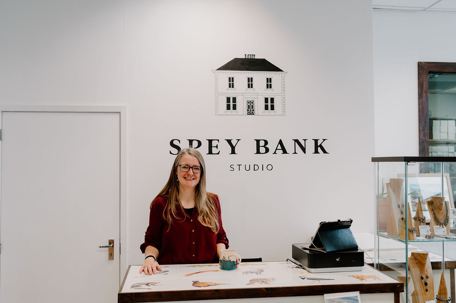 A woman stands smiling behind the Spey Bank Studio counter, small prints and cards spread across it beneath a painted house logo.