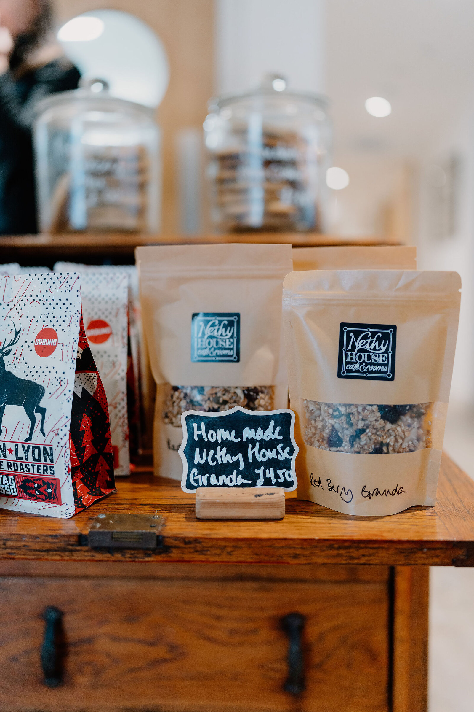 Bags of homemade Nethy House granola stand on a wooden counter beside a chalkboard price tag and jars of biscuits.