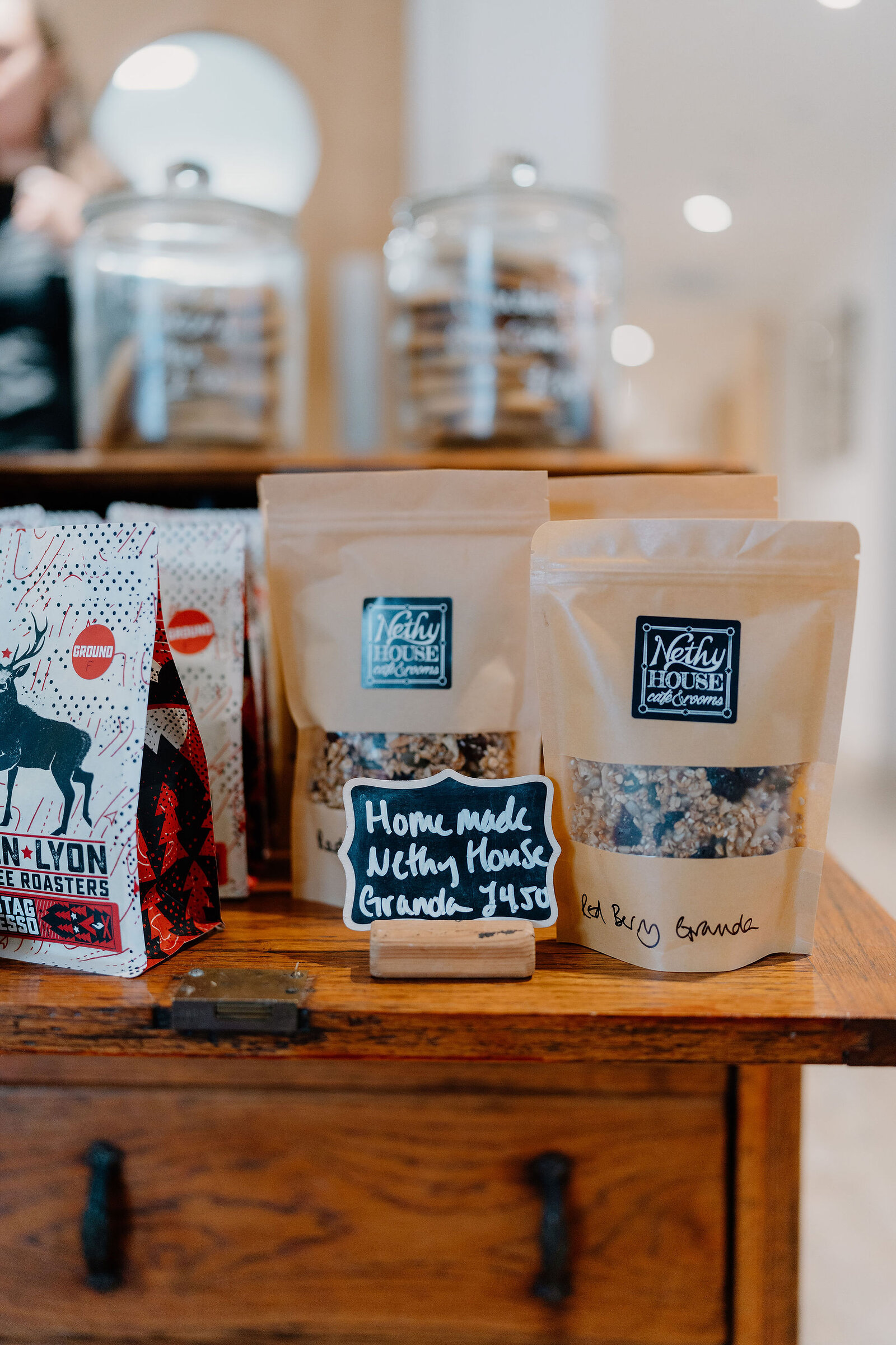 Bags of homemade Nethy House granola stand on a wooden counter beside a chalkboard price tag and jars of biscuits.