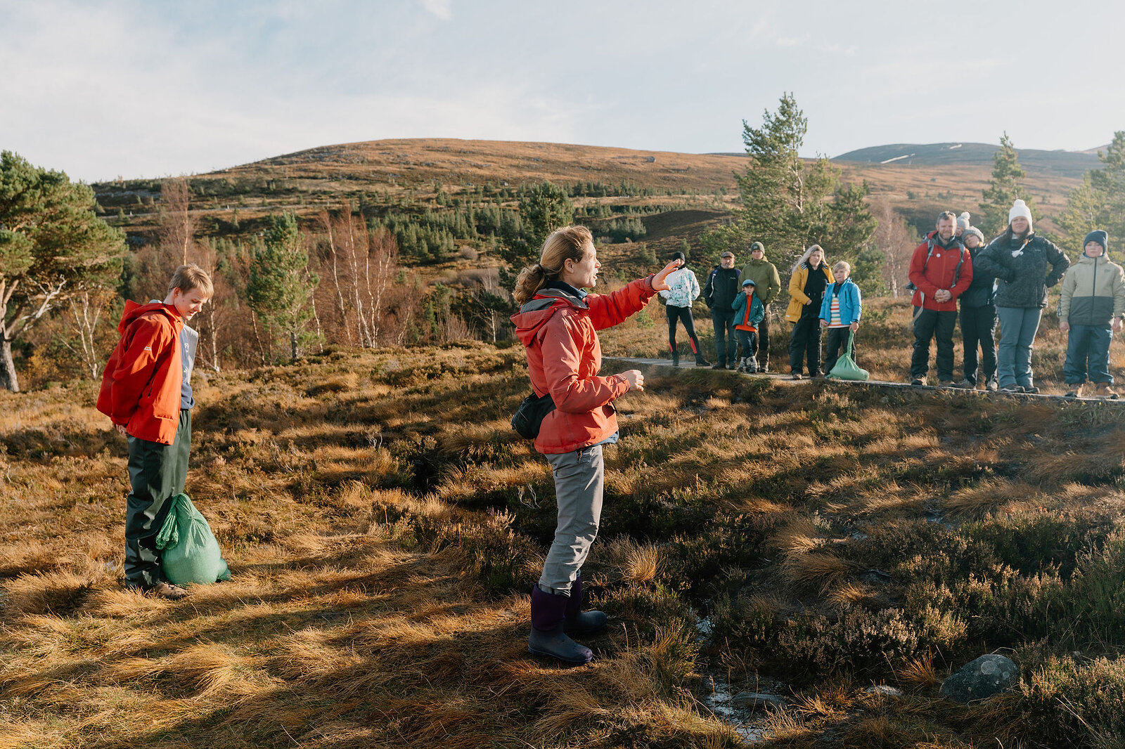 A guide in a red jacket gestures to a small group gathered on a heather moorland hillside, forestry and rolling hills stretching behind them.