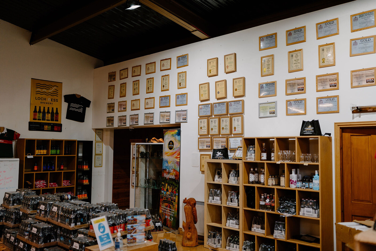Rows of framed awards line a white wall above shelves of bottled beer either side of a doorway, a carved wooden figure standing near the entrance.