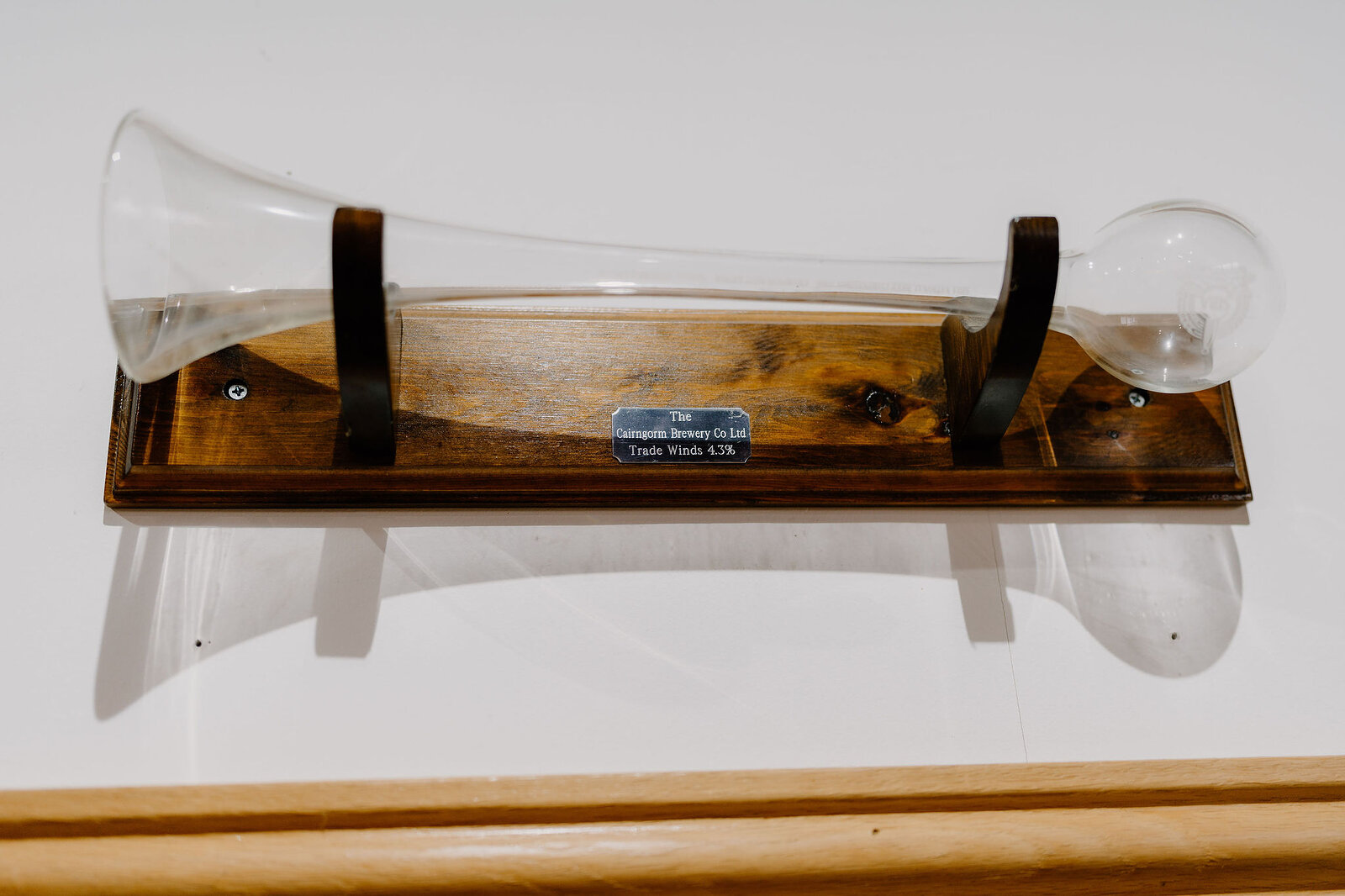 An antique glass yard-of-ale vessel stands mounted on a wooden base, a brass plaque naming the Cairngorm Brewery fixed beneath it.