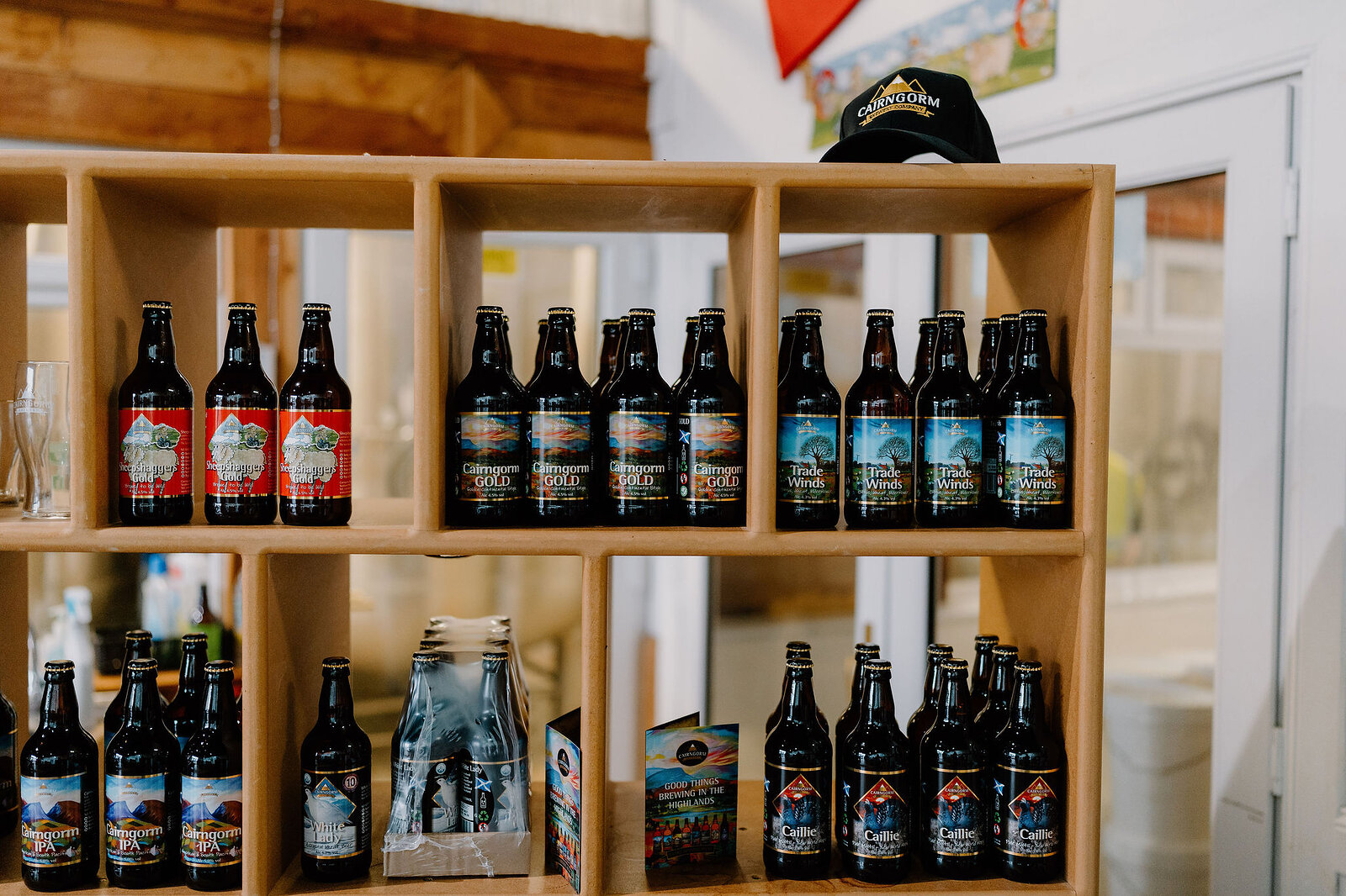 Shelves of bottled beer with printed labels stand beneath a branded cap hanging from the shelf edge, a glass door reflecting light beside them.