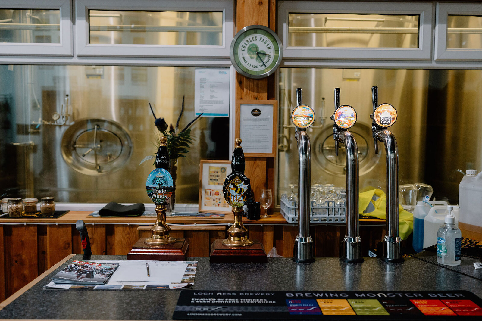 A brewery bar counter holds beer taps and a hand pump beneath a wall clock, pamphlets and a brewing poster laid out beside a bottle of hand sanitiser.