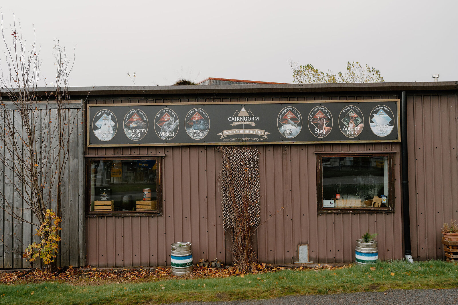 A brewery building's exterior carries a row of circular product logos along its roofline, two barrels standing either side of the door between small display windows.