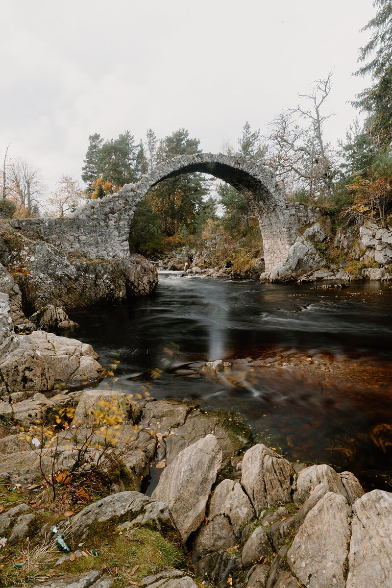 A stone humpback bridge over a river captured in a long exposure, the water blurred silk-smooth and tinted amber where fallen leaves drift beneath the arch.