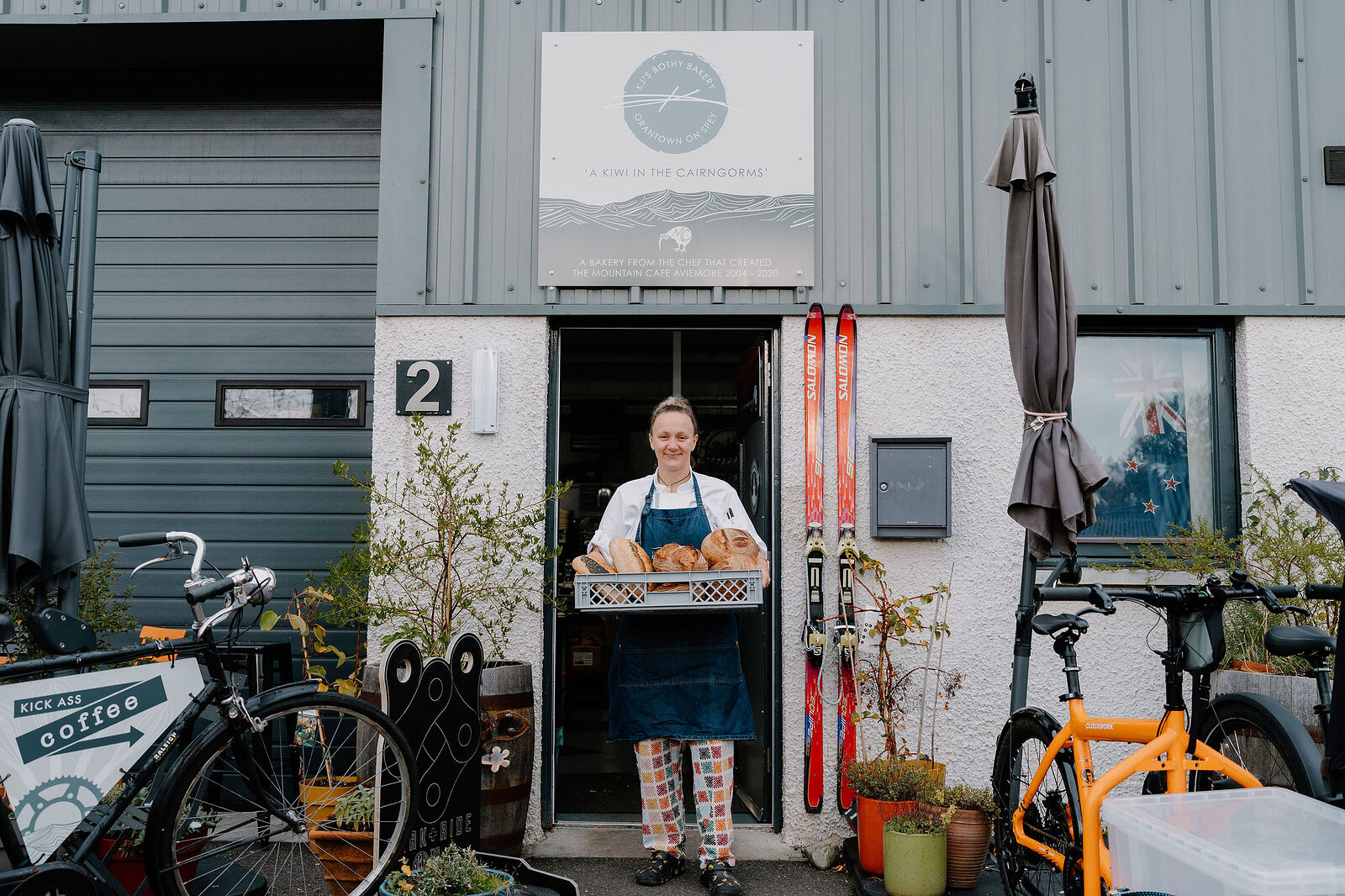 A woman in an apron holds a crate of bread at the doorway of KJ's Bothy Bakery, bicycles and folded umbrellas standing outside beneath the shop sign.