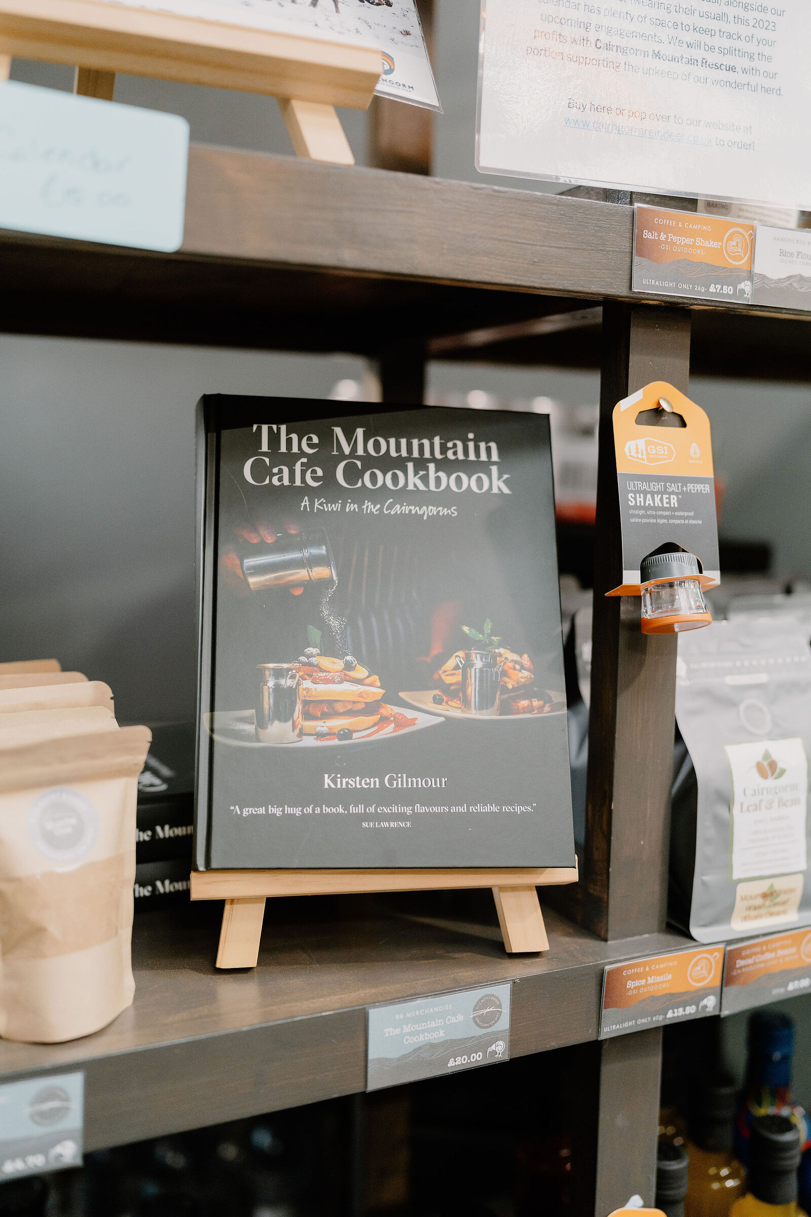 The Mountain Cafe Cookbook, A Kiwi in the Cairngorms by Kirsten Gilmour stands displayed on a small easel among shop shelving and price tags.
