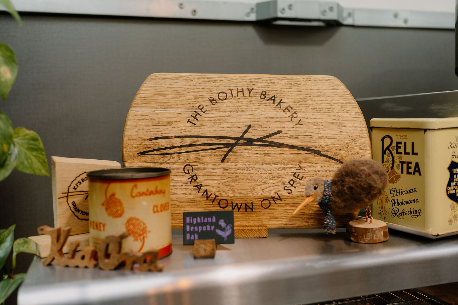 A carved board reading The Bothy Bakery, Grantown on Spey stands among tins, a felt kiwi bird ornament and a Highland Bespoke tag on a shelf.