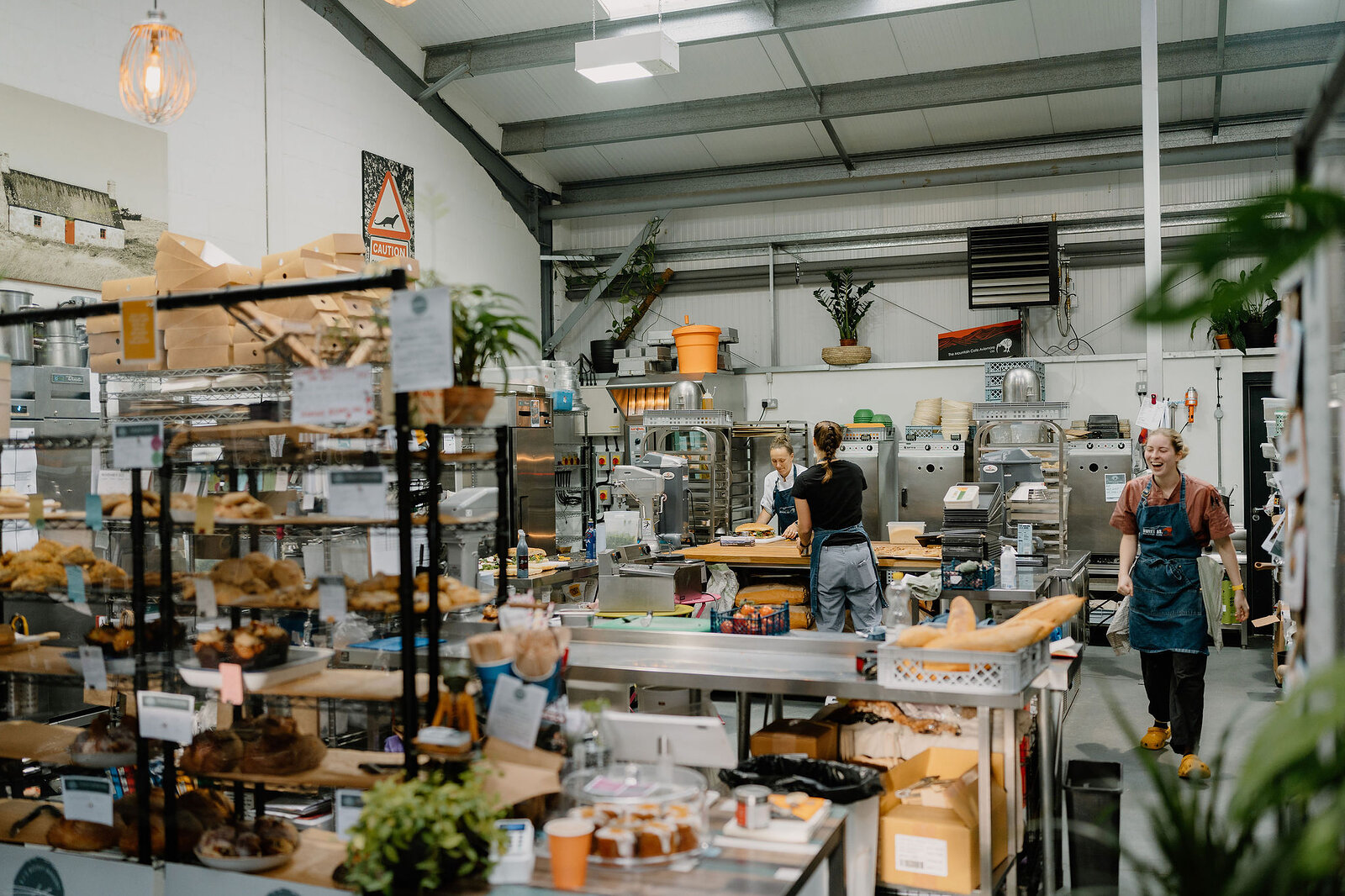 Two staff work among shelving and equipment in a bakery, trays of pastries and loaves stacked on racks around them.