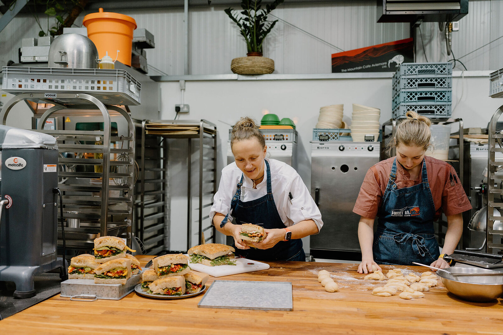 Two women prepare sandwiches at a wooden table in a bakery kitchen, filled rolls laid out on a tray in front of them.