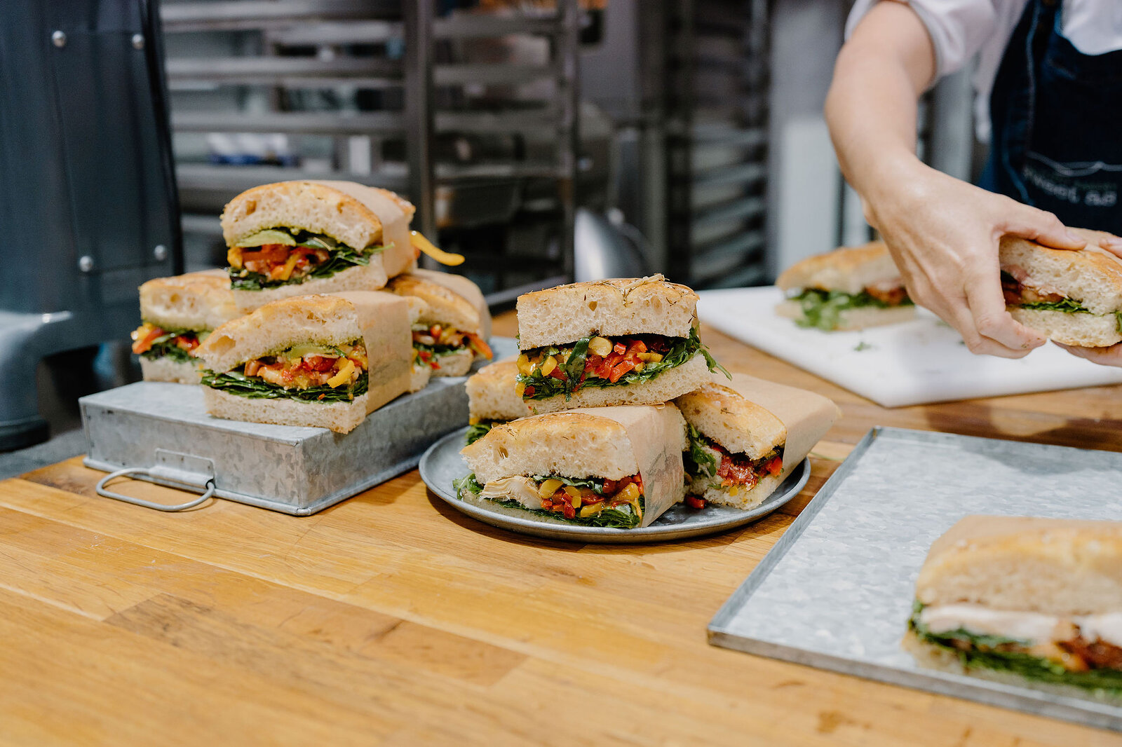 Stacked sandwich halves filled with salad and vegetables are piled on stone boards and plates, a hand placing another sandwich on a tray behind.