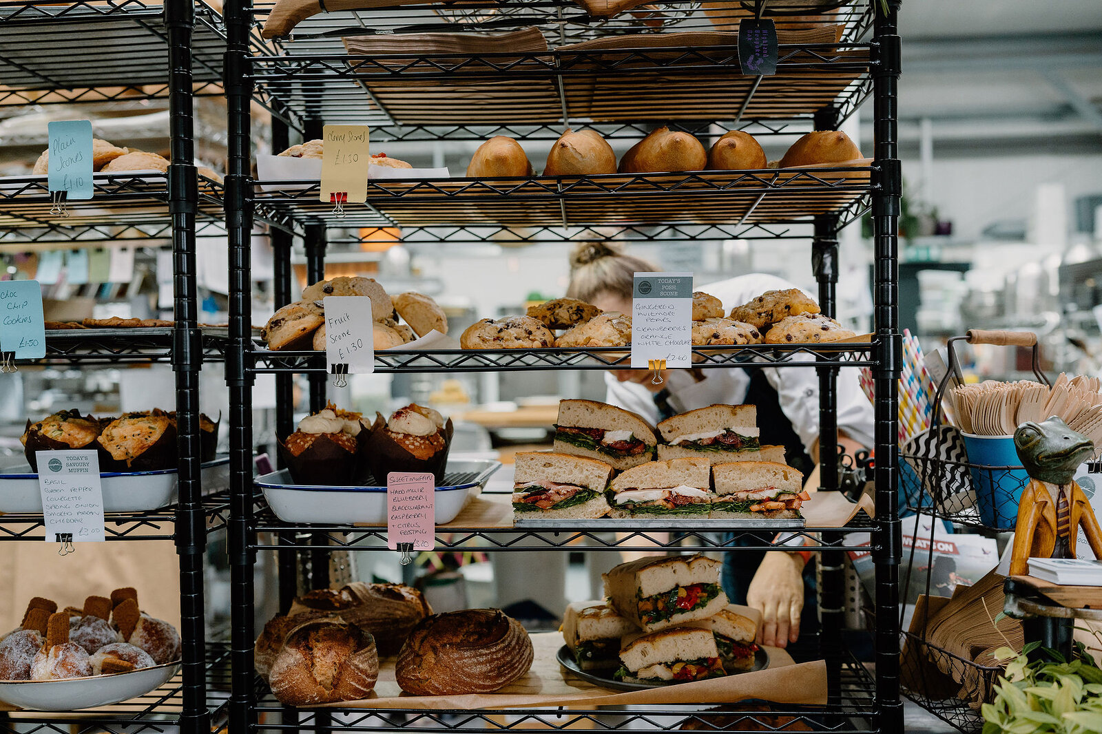 Tiered wire shelving holds loaves, pastries and sandwiches with handwritten price signs, a staff member working behind the display.