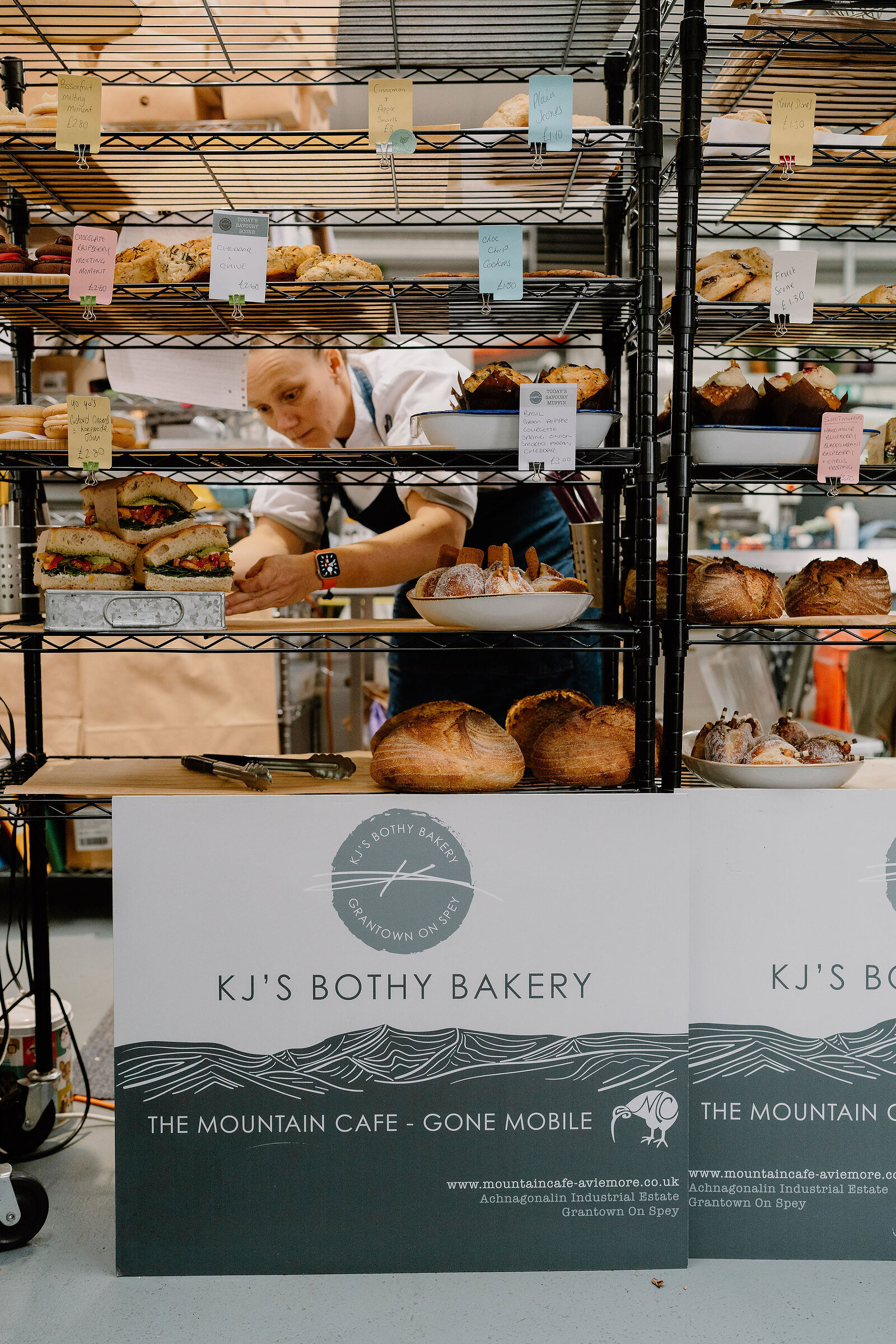 Sign boards reading KJ's Bothy Bakery, The Mountain Cafe Gone Mobile, stand before tiered shelving of loaves and sandwiches, a baker working behind.