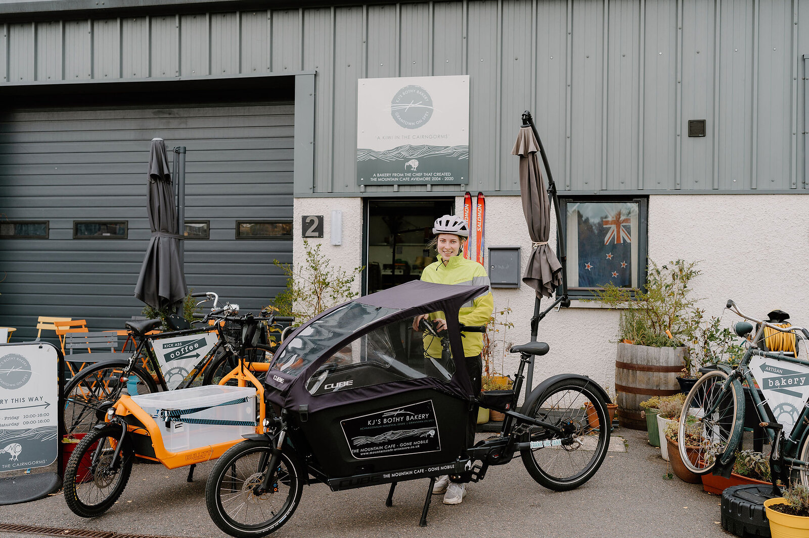 A woman in a cycling helmet sits on a cargo bike outside a workshop unit, folded umbrellas and a bicycle standing against the wall behind.