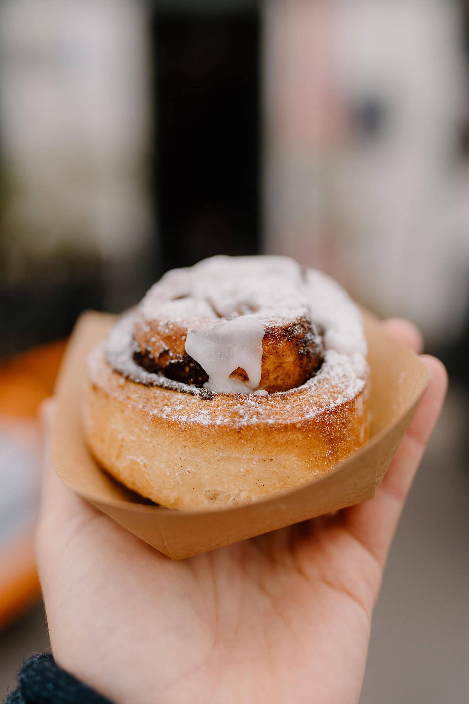 A hand holds a cinnamon roll dusted with icing sugar and drizzled with white icing, steam rising faintly from its centre.