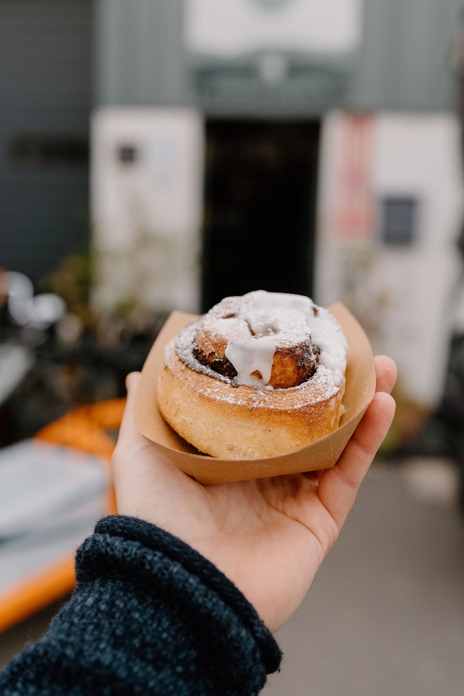 A hand holds a cinnamon roll dusted with icing sugar and drizzled with white icing, a shop doorway blurred behind.