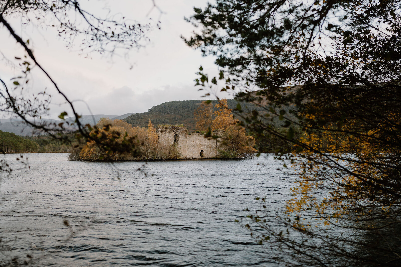 A ruined castle on a small island is glimpsed through autumn branches, hills rising beyond the still loch.