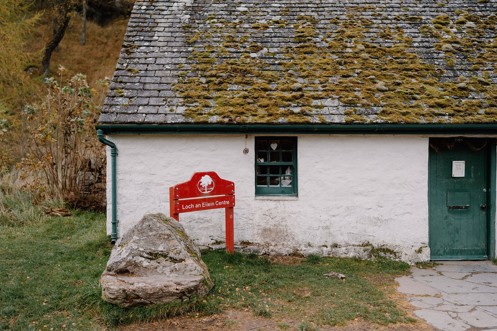 A red sign reading Loch An Eilein Centre hangs on a whitewashed cottage with a mossy slate roof, a large boulder resting on the grass in front.