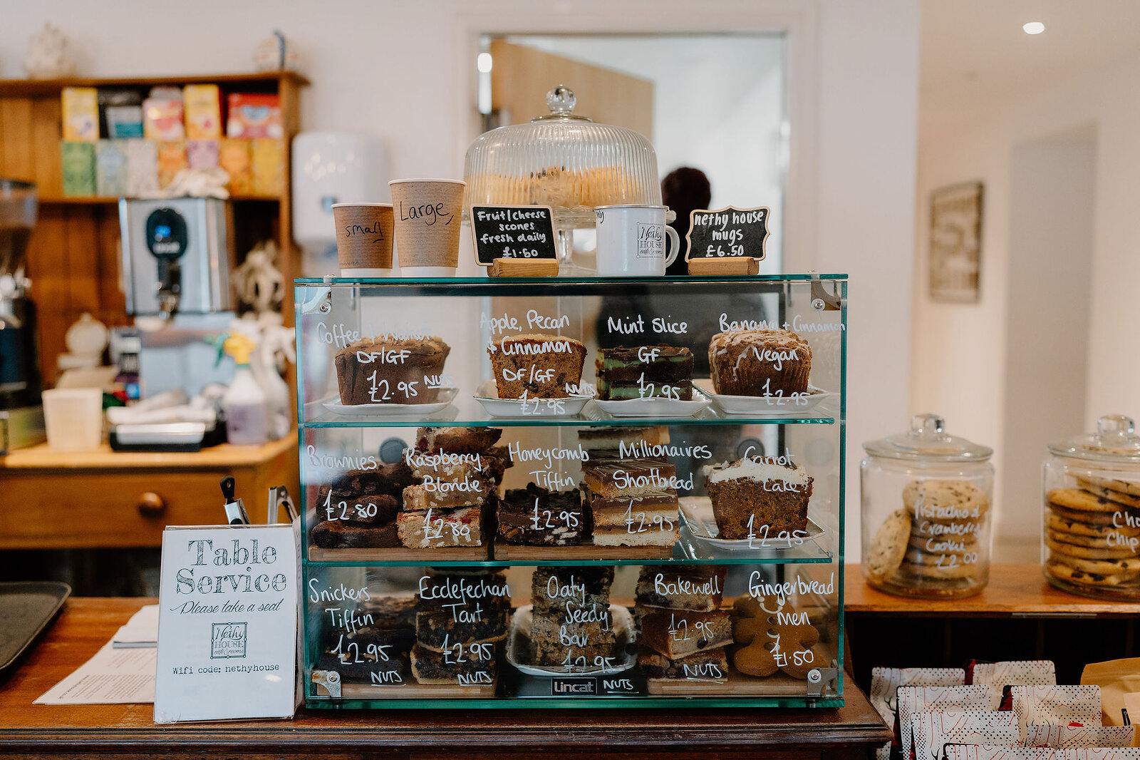 A glass display case of tray bakes and muffins sits on a cafe counter with chalkboard price labels, a Table Service sign and jars of biscuits nearby.