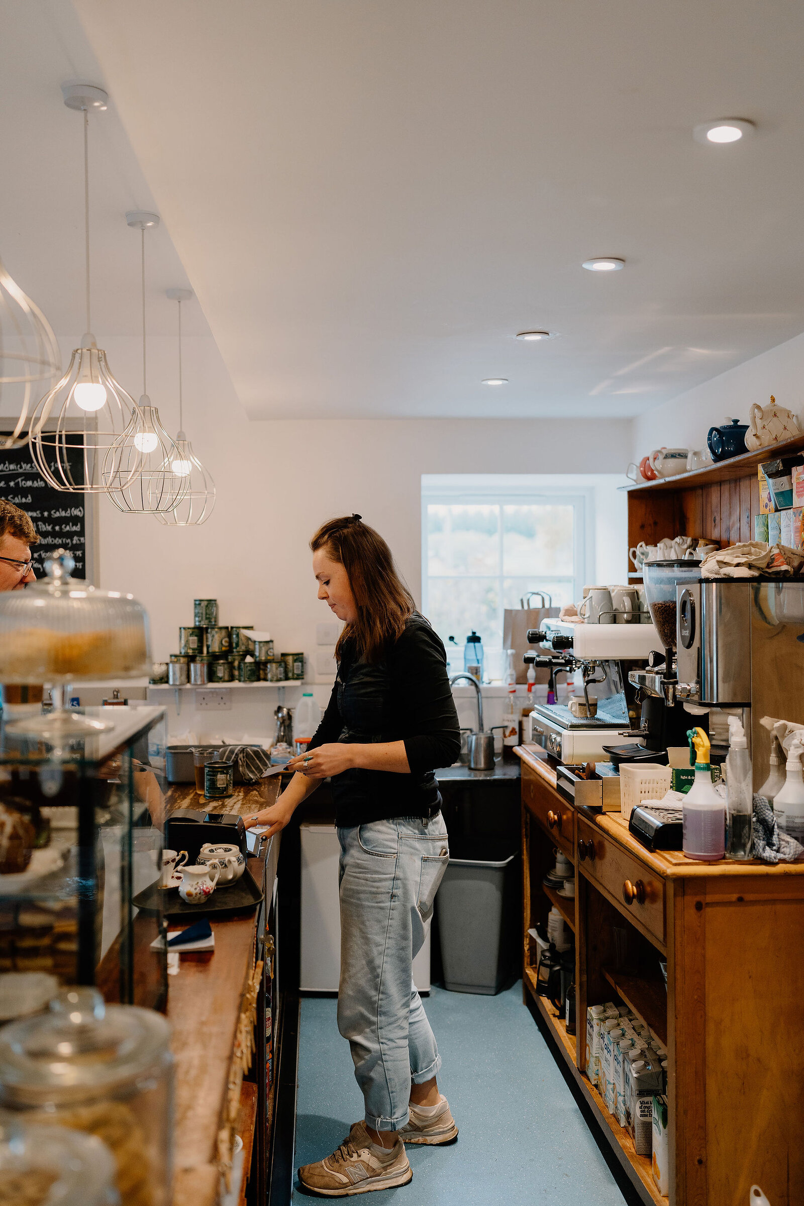 A woman works at a counter in a tearoom shop, pendant lights hanging above shelves of jars, tins and packaged goods.