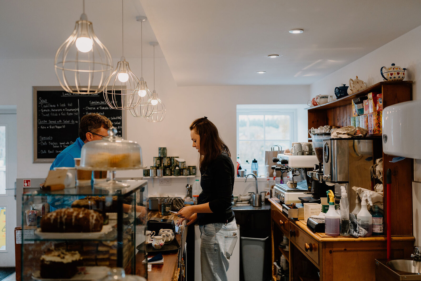 A woman serves at a tearoom counter while a man browses nearby, pendant lights hanging above a glass cake display.
