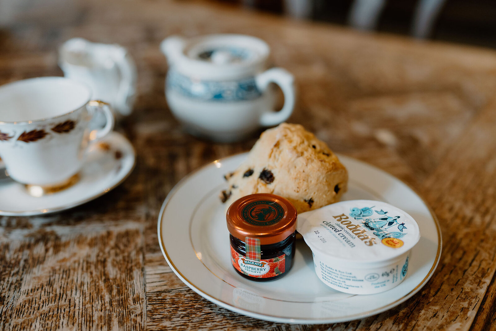A fruit scone, a jar of jam and a tub of clotted cream sit on a plate beside a floral teacup and a milk jug on a wooden table.
