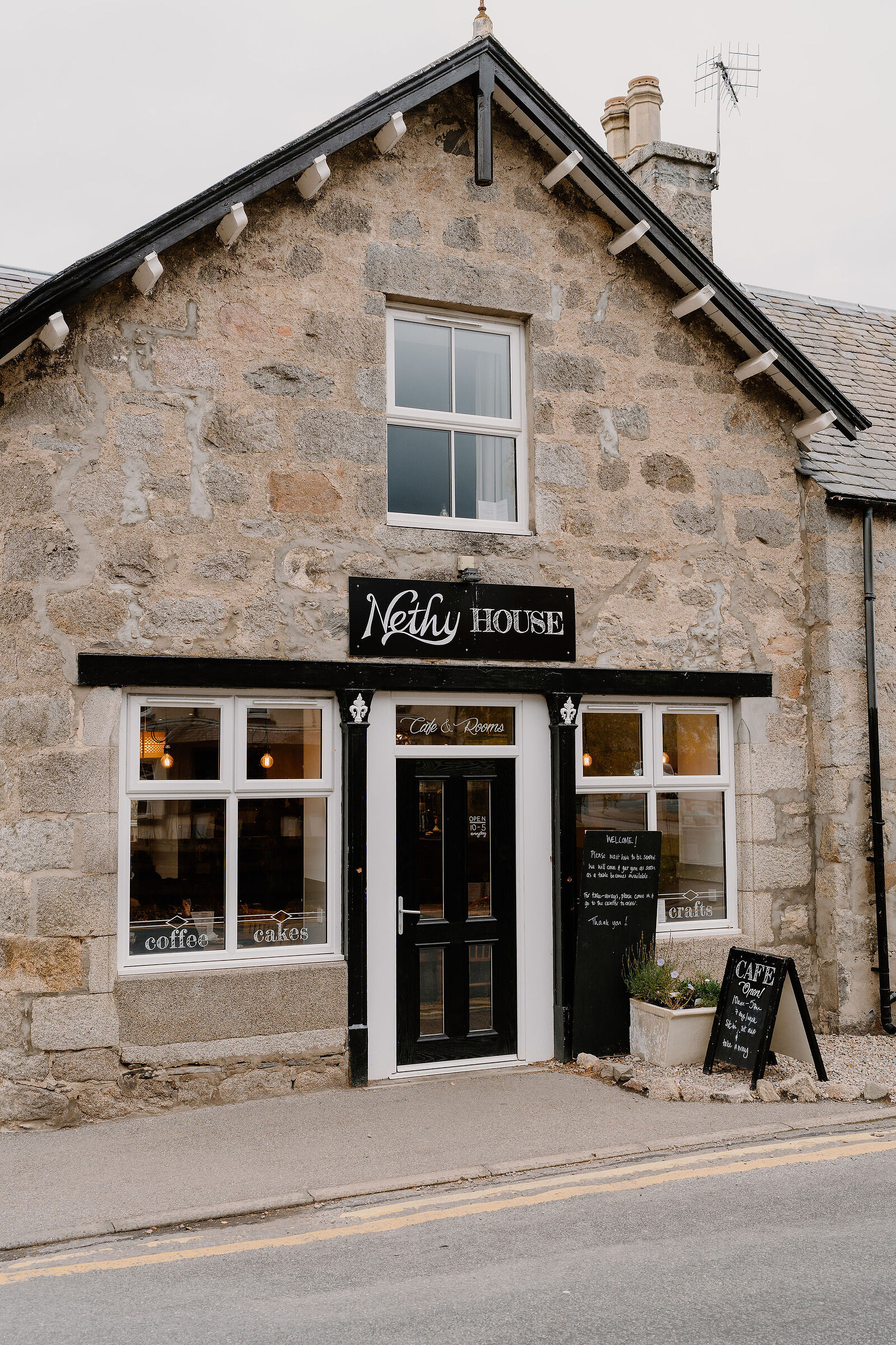 A stone corner building signed Nethy House, Cafe and Rooms, holds a bakery and cafe behind black-framed windows, chalkboard signs and potted plants at the door.