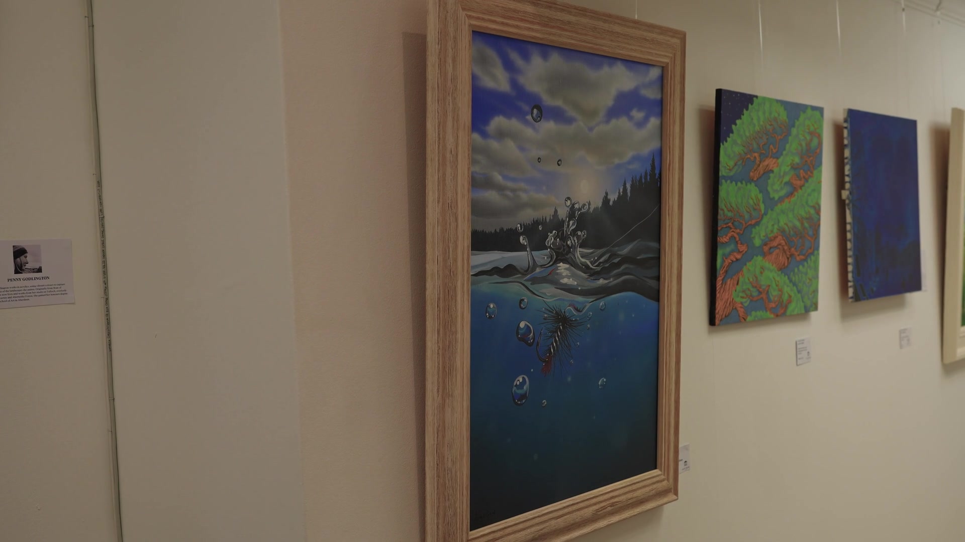 A framed acrylic painting of a moonlit loch and fishing fly hangs beside smaller canvases in a gallery, an artist's biography card mounted on the wall.