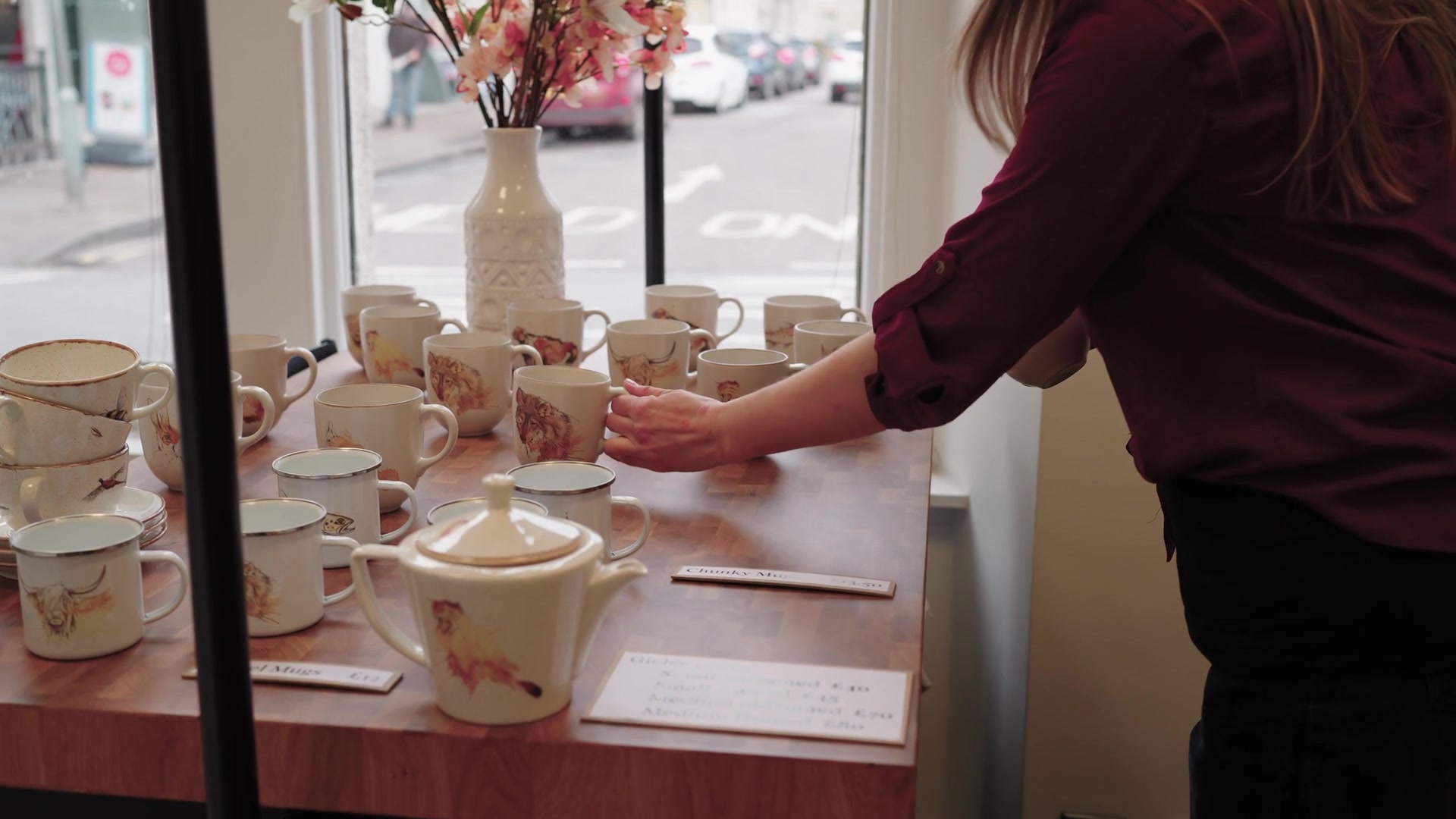 A woman reaches for an animal-print mug on a table of enamel mugs and teapots, a "Chunky Mugs" price sign among them and a street visible through the window behind.