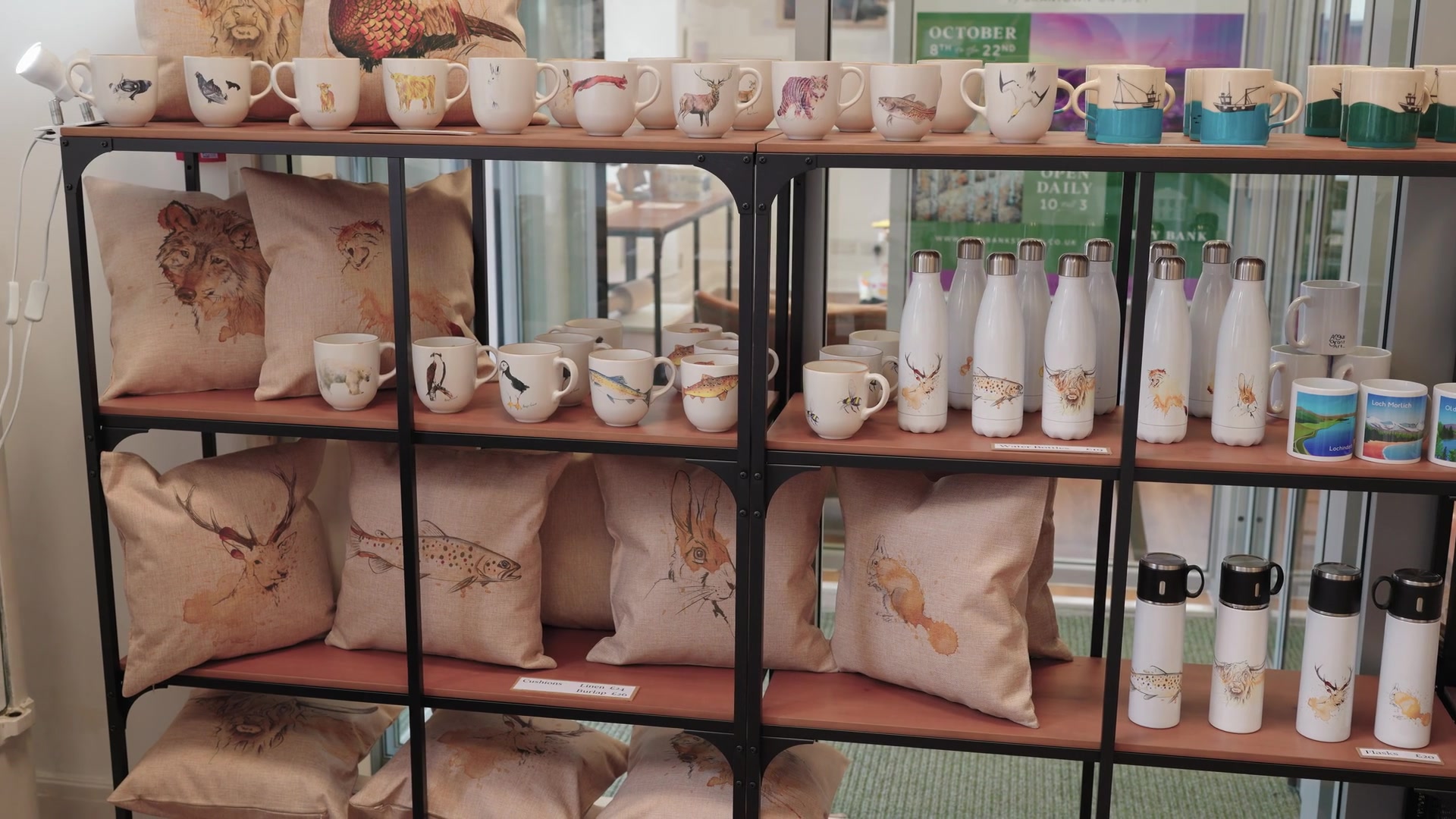 A shelf of animal-print mugs, cushions and water bottles fills a gift shop display beneath a sign advertising an October event.