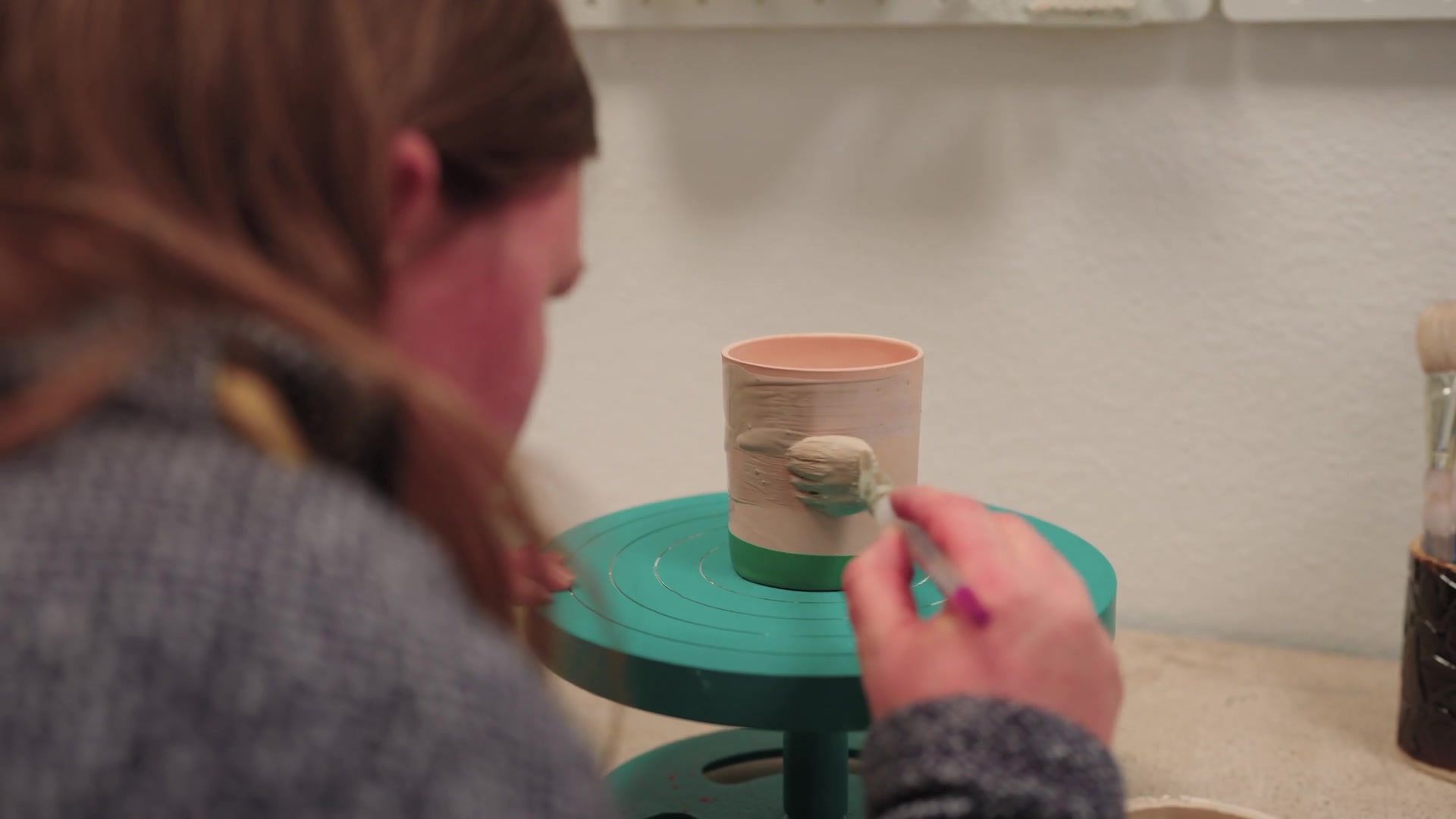 A woman shapes a small clay bowl at a pottery wheel, water splashing over her hands as the clay spins.