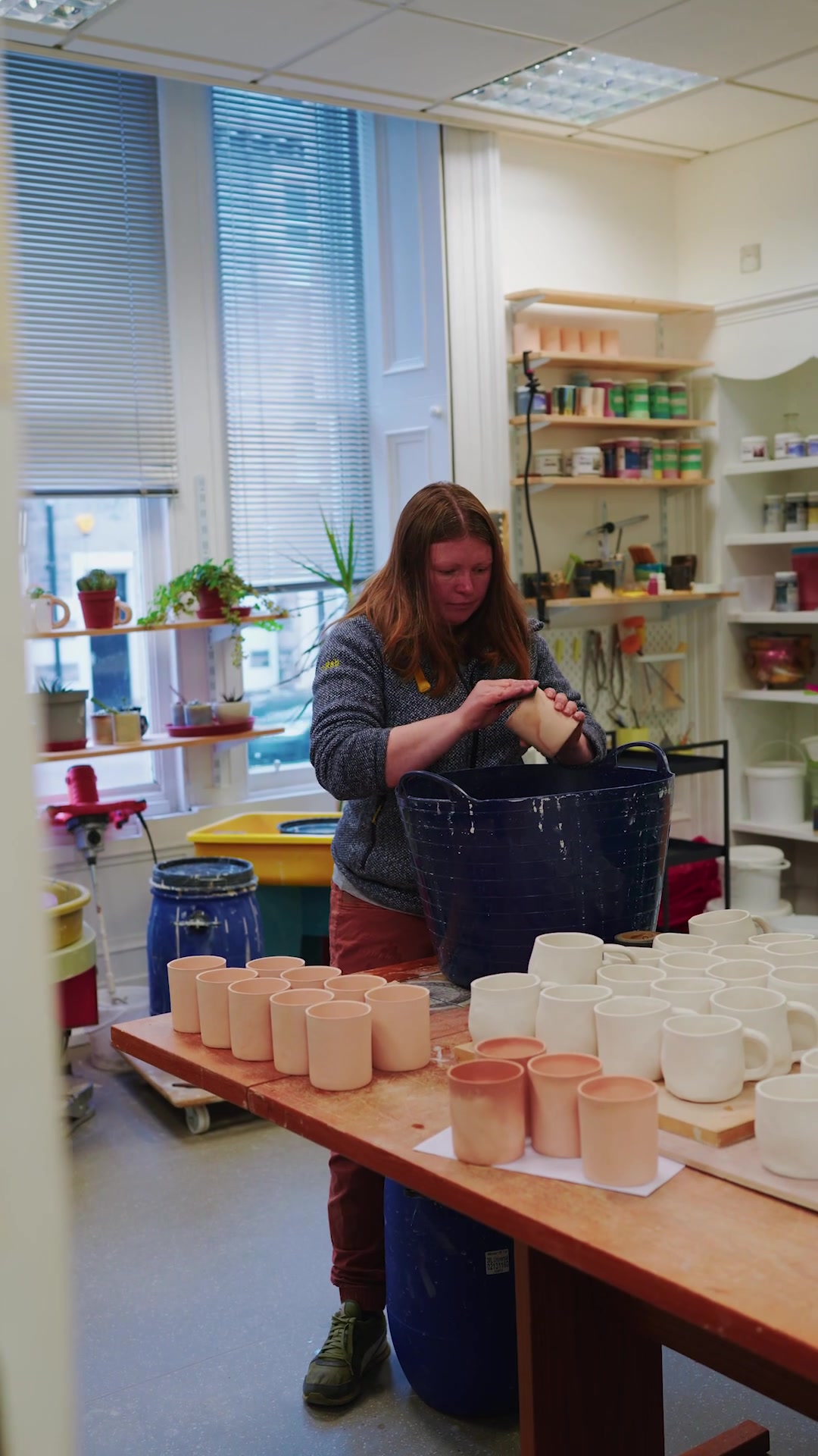 A woman shapes the rim of an unfired mug over a blue tub, rows of pale clay mugs and glazed white ones lined up on the workbench beside her.