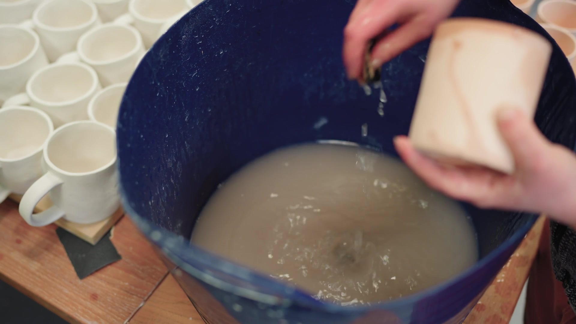 Hands dip a pale mug into a bucket of blue glaze, rows of finished white mugs lined up on the table behind.