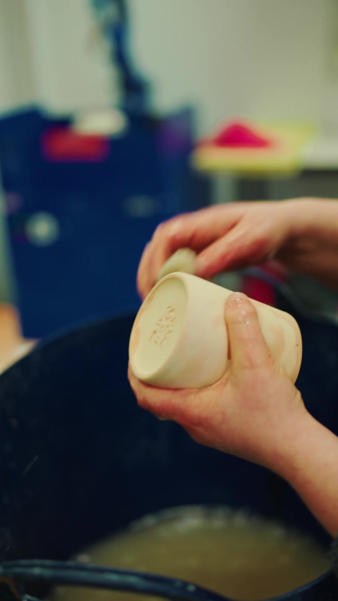A potter's clay-flecked hands turn a small stoneware cup, trimming its rim over a work bowl.