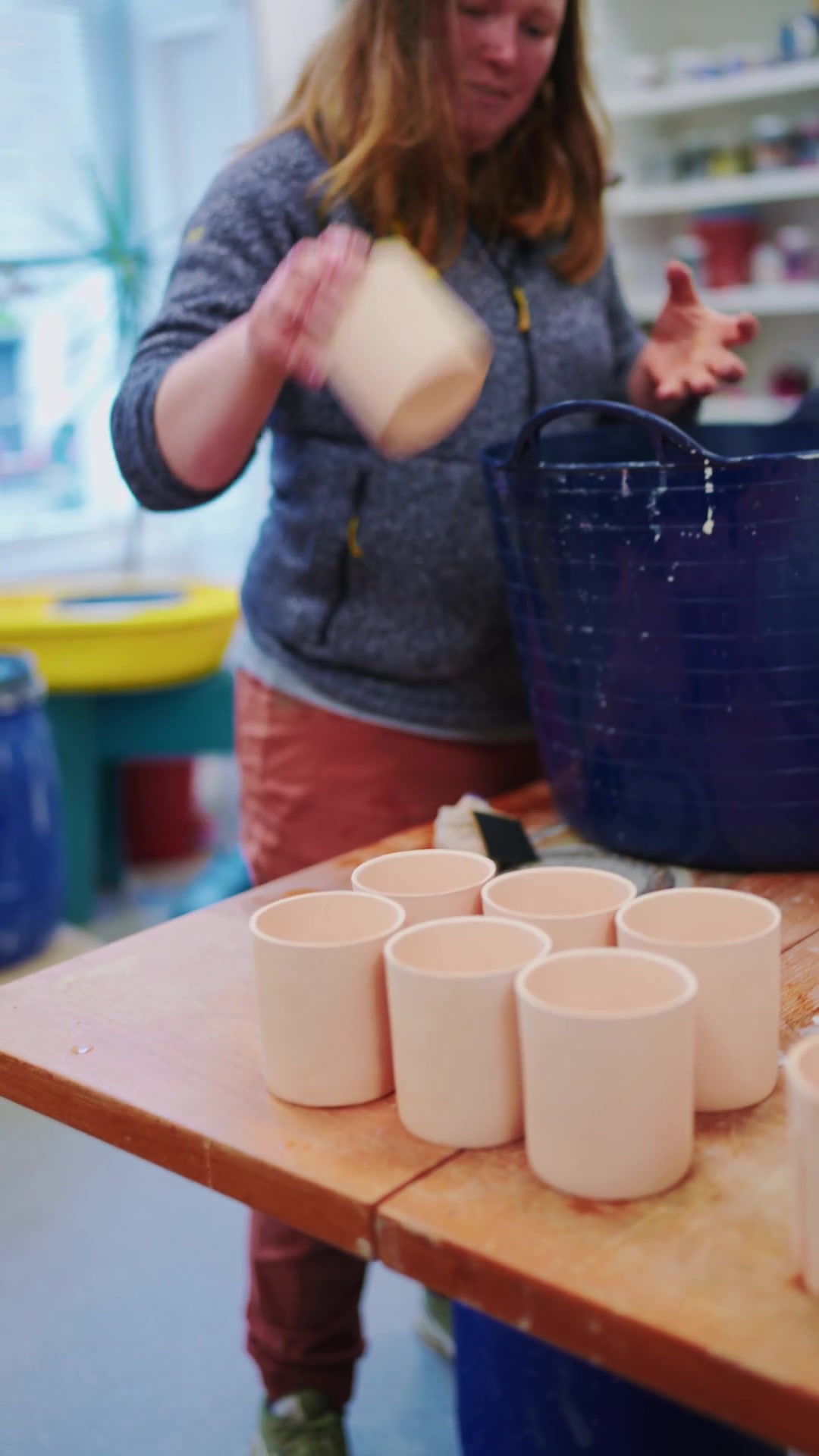 A woman lifts a pale stoneware cup from a row of identical cups on a workbench, a blue bucket and shelves of glazes behind her.