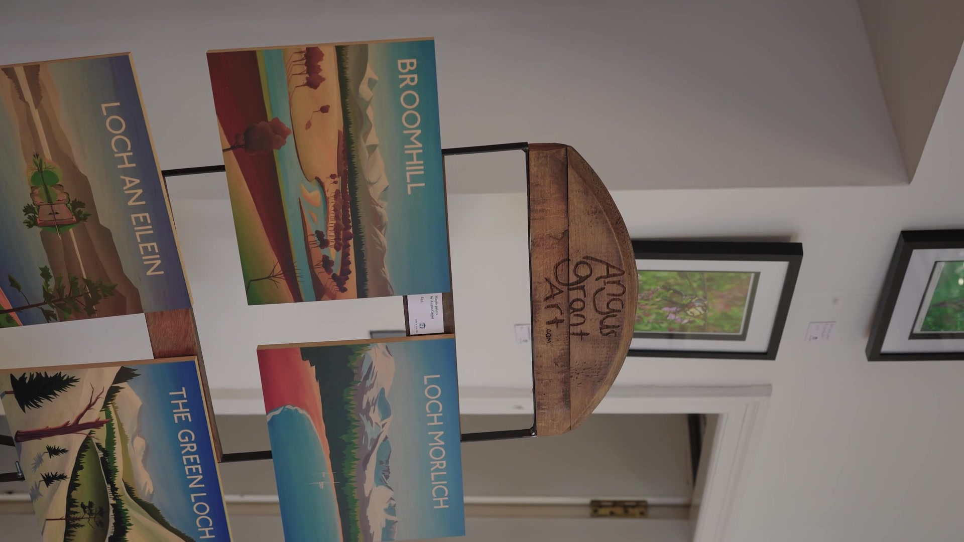 Illustrated prints titled "Loch an Eilein", "Broomhill" and "Loch Morlich" hang from a rack beside a wooden "Angus Grant Art" sign.