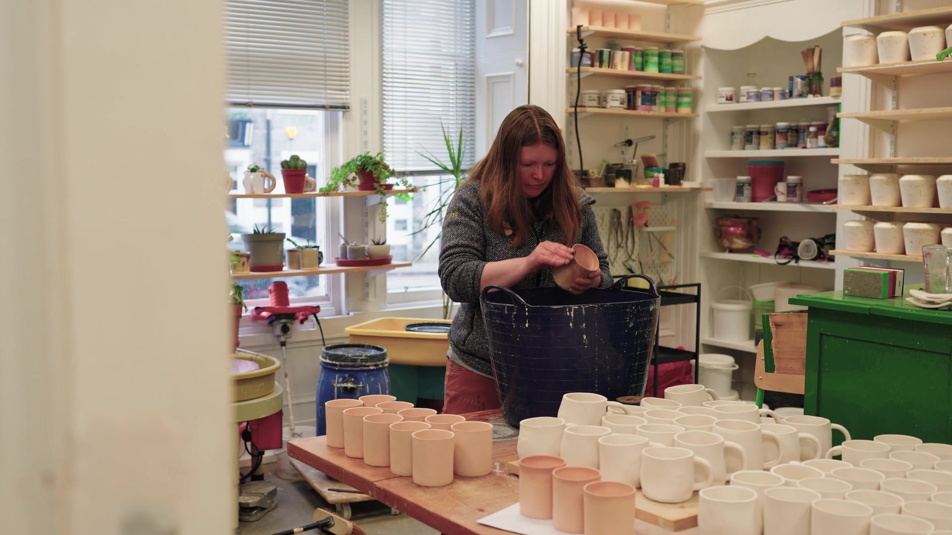 A woman lifts a pale stoneware cup from a blue tub, rows of unglazed and white-glazed mugs lined up on a workbench before her.