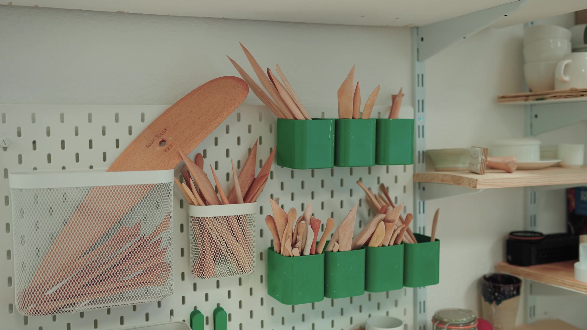 Wooden pottery tools stand bristling from green wall-mounted pots on a pegboard, glazed mugs stacked on a shelf beside them.