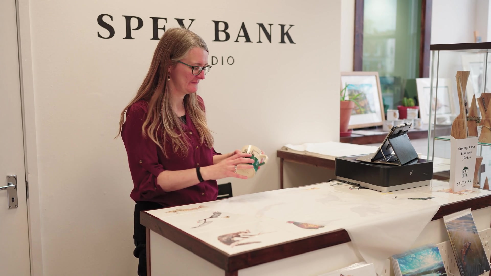 A woman in glasses examines a hand-painted mug at a till counter lettered "Spey Bank Studio", framed wildlife illustrations and greetings cards displayed around her.