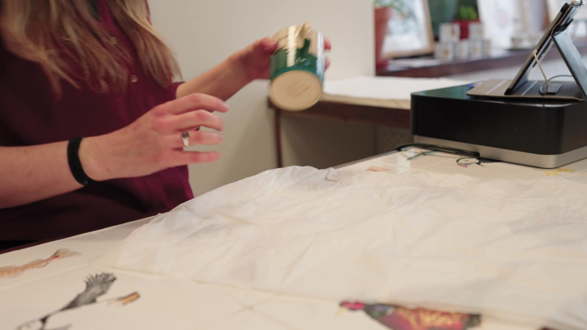A woman's hands wrap a hand-painted mug in tissue paper on a counter scattered with illustrated bird cards, a card reader and picture frames visible behind her.