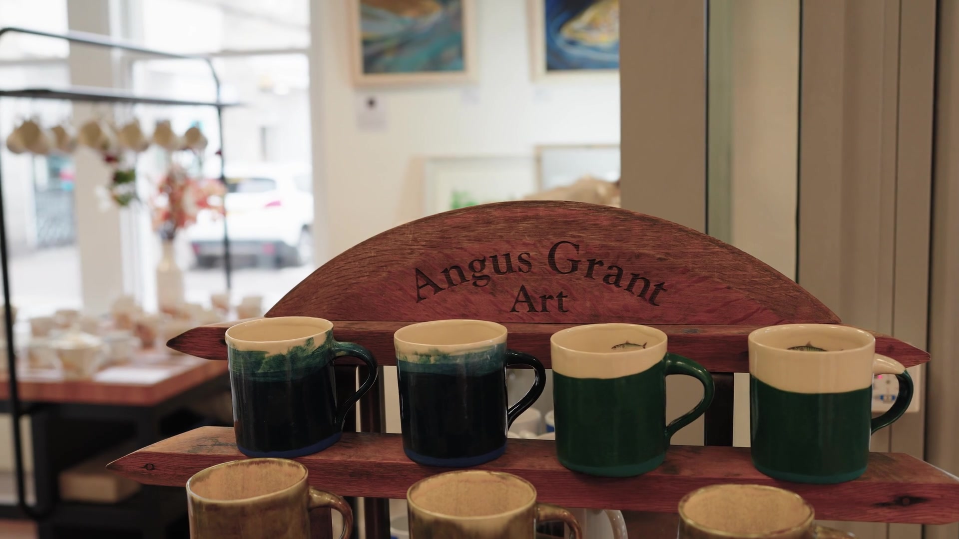 Green and cream glazed mugs, one printed with a fish, are lined up on a rack beneath a wooden "Angus Grant Art" sign, framed seascape paintings hanging on the gallery wall behind.