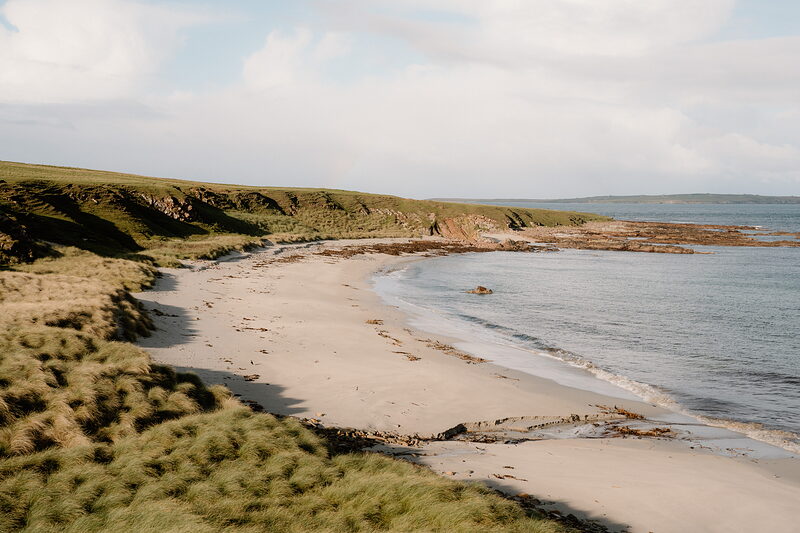 An aerial view of a curving sandy beach lit by low evening light, weathered red rock at the shoreline and dune grass rising to the clifftop.
