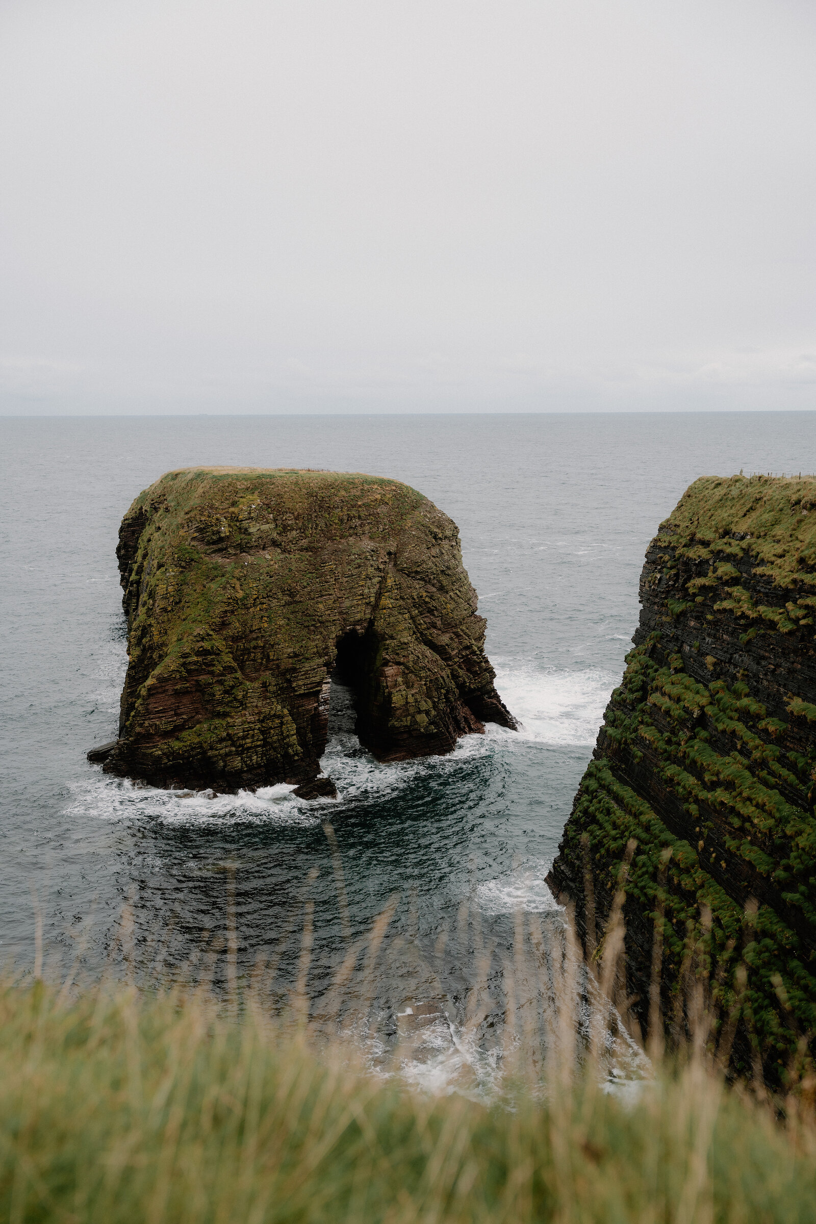 A rounded sea stack pierced by a low arch stands offshore, waves breaking through the gap at its base.