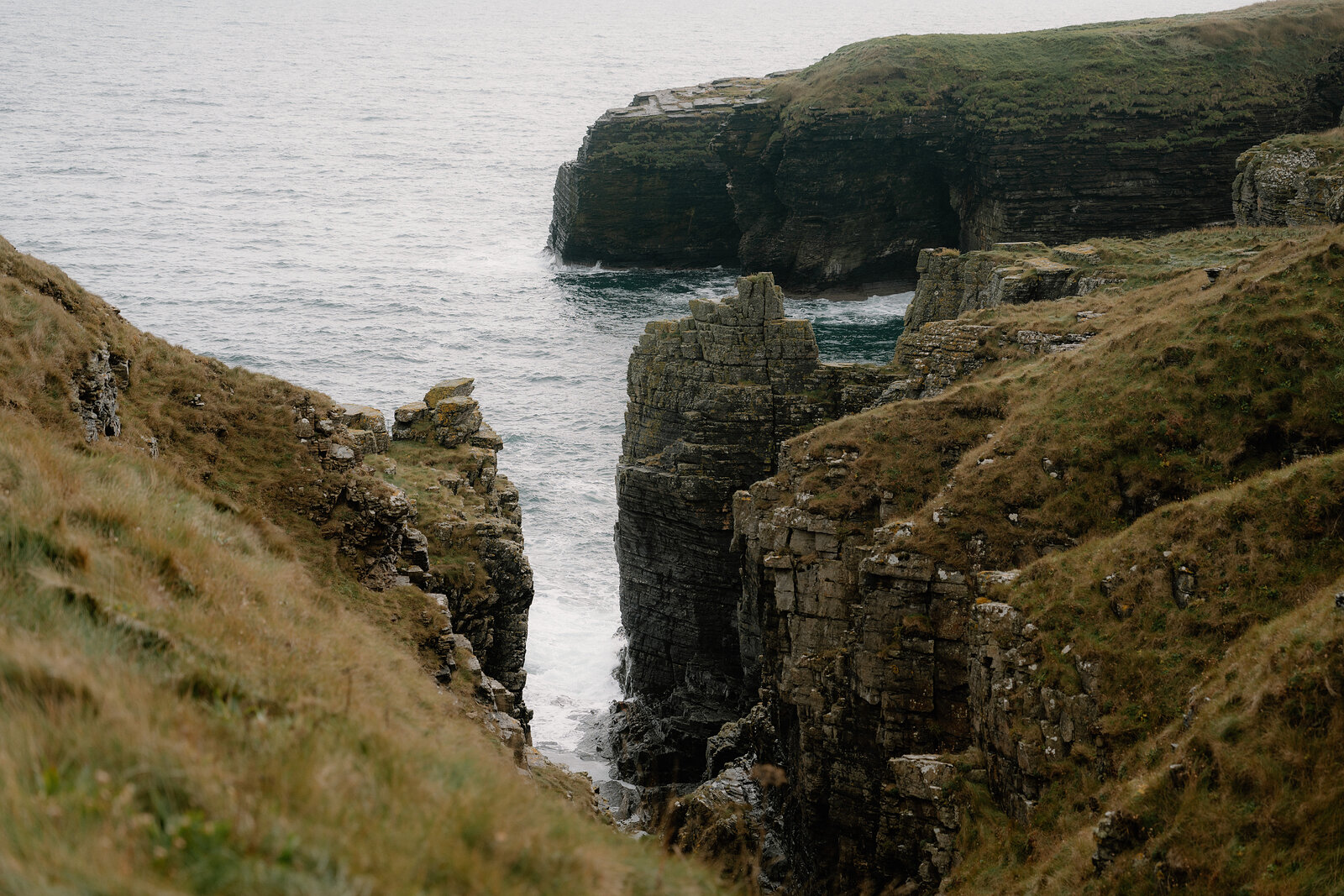 Waves crash into a narrow sea inlet between steep grassy cliffs and layered rock walls.