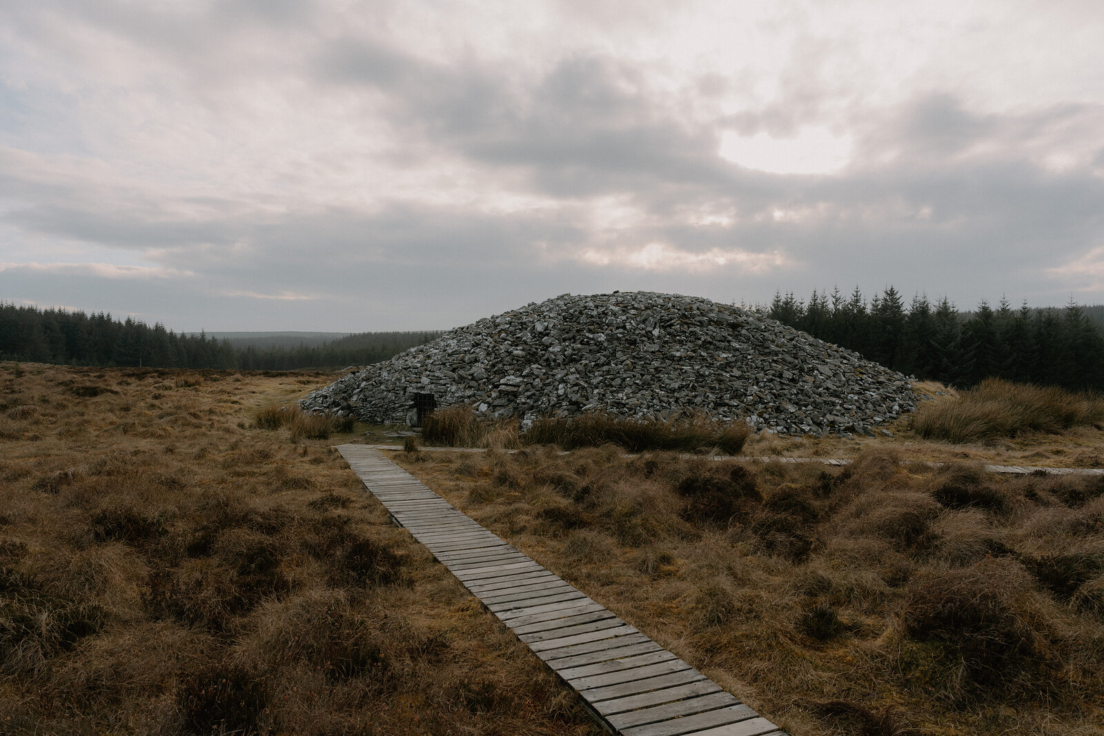 A wooden boardwalk leads across moorland to a large chambered cairn built of stacked stone, forest lining the horizon.