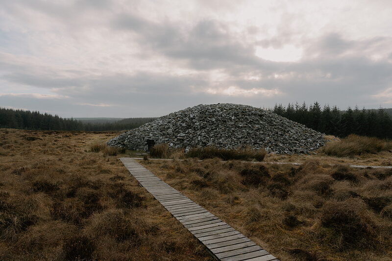 A wooden boardwalk leads across moorland to a large chambered cairn built of stacked stone, forest lining the horizon.